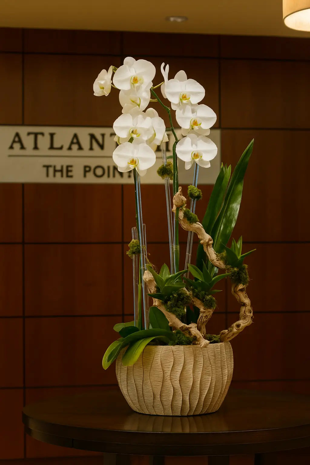 A floral arrangement of white orchids in an elegant round vase with decorative branches and green accents, placed in a modern interior with wood and glass elements.