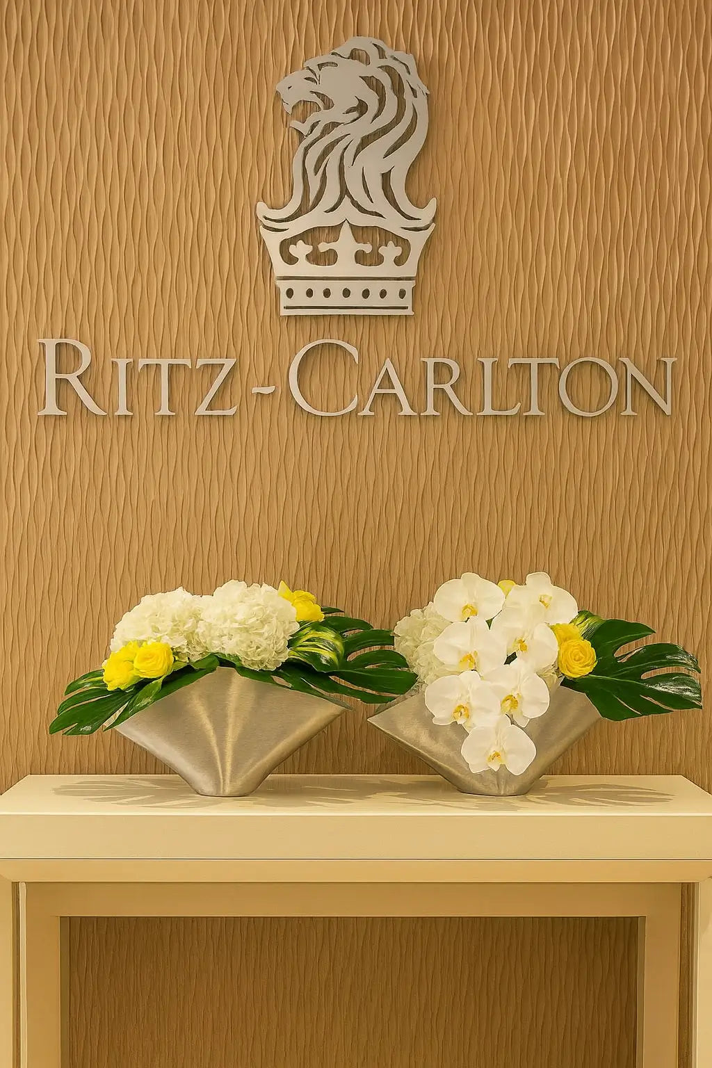 A premium interior view of The Ritz-Carlton Residences, Sunny Isles Beach. The iconic Ritz-Carlton crown logo is displayed on a textured wall, with elegant floral arrangements of white and yellow flowers placed in modern metallic vases on a sleek console table, creating a refined and luxurious atmosphere.