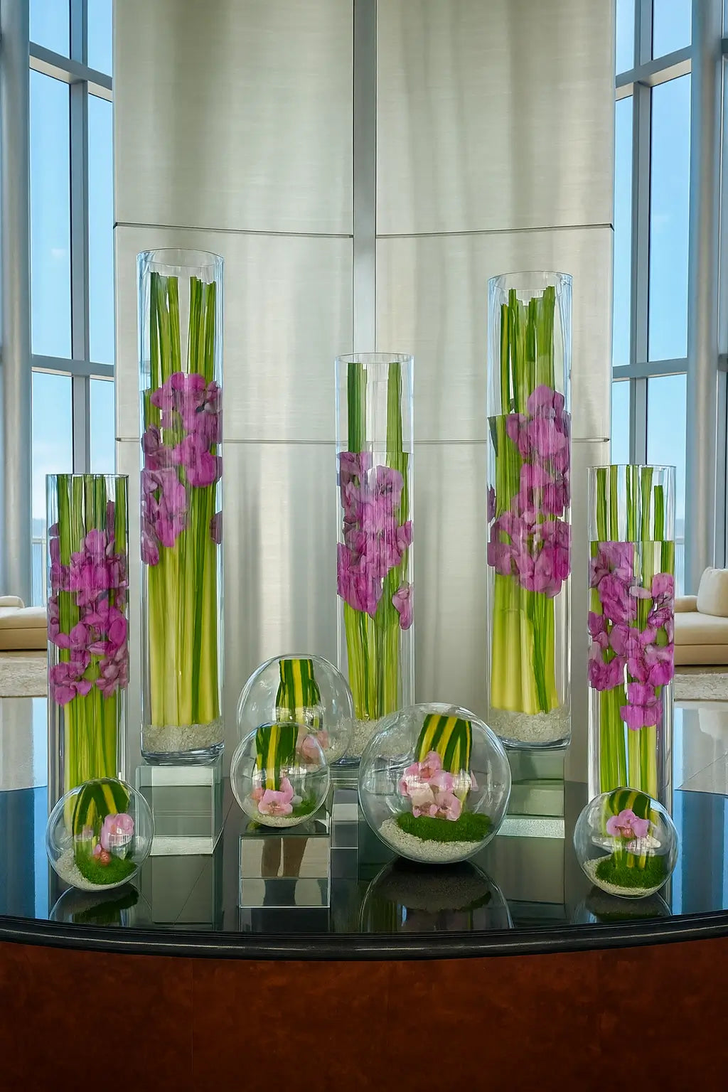 A floral arrangement of five tall glass vases with decorative greenery and white orchids, displayed in a contemporary lobby interior with large windows and modern seating in the background.