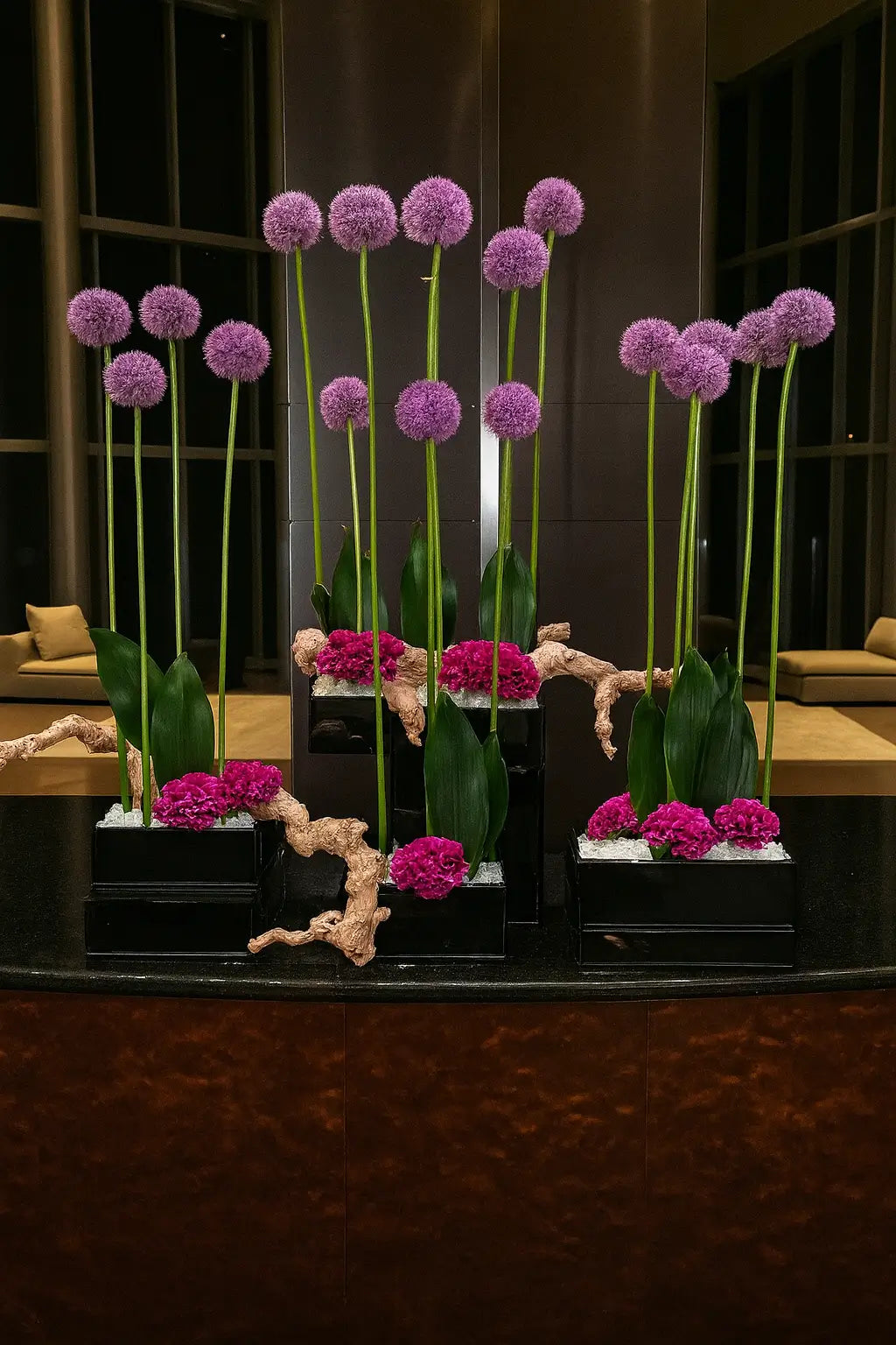 A digital photograph features multiple floral arrangements with tall purple allium flowers and accent greenery, displayed in sleek black rectangular vases on a glossy round table, set in a contemporary lobby with warm lighting and modern seating.