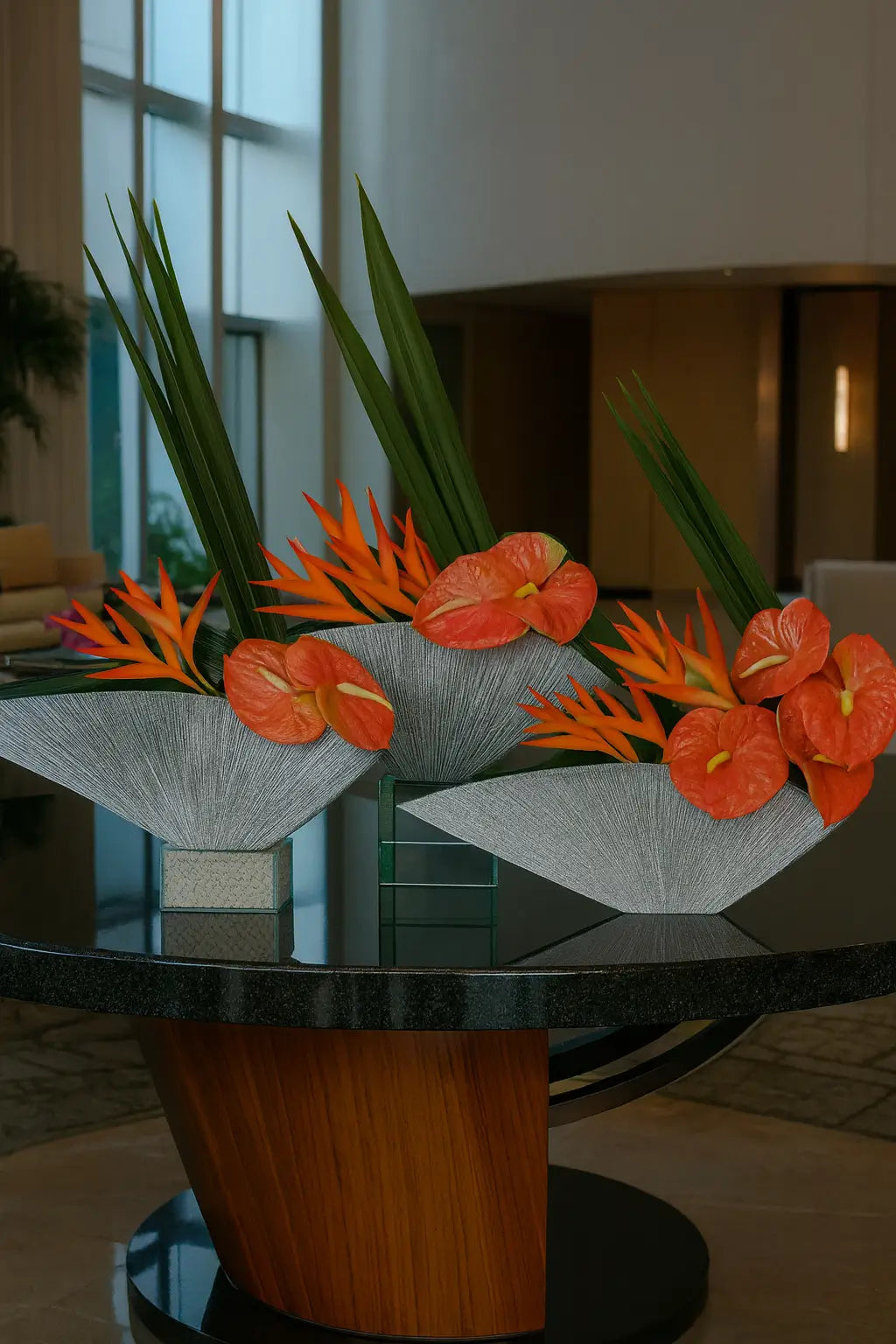 A digital photograph features three elegant floral arrangements displayed in silver textured vases, each filled with lush green leaves, orange bird-of-paradise flowers, and clusters of pink anthuriums. The compositions are arranged symmetrically on a reflective glass surface, set against a contemporary lobby with soft beige seating and large floor-to-ceiling windows that allow natural light to brighten the space.