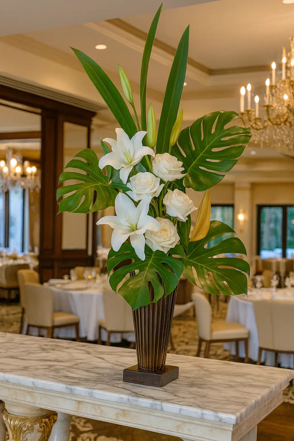 Luxury tropical floral arrangement with white lilies, roses, and monstera leaves in a tall vase, set on a marble table in an elegant fine dining restaurant.