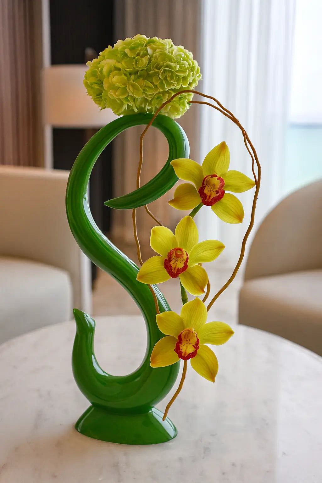 Elegant floral arrangement in the style of Turnberry Ocean Club Residences: a green curved vase echoing the shape of the logo, with yellow orchids and green hydrangea, complemented by decorative branches.