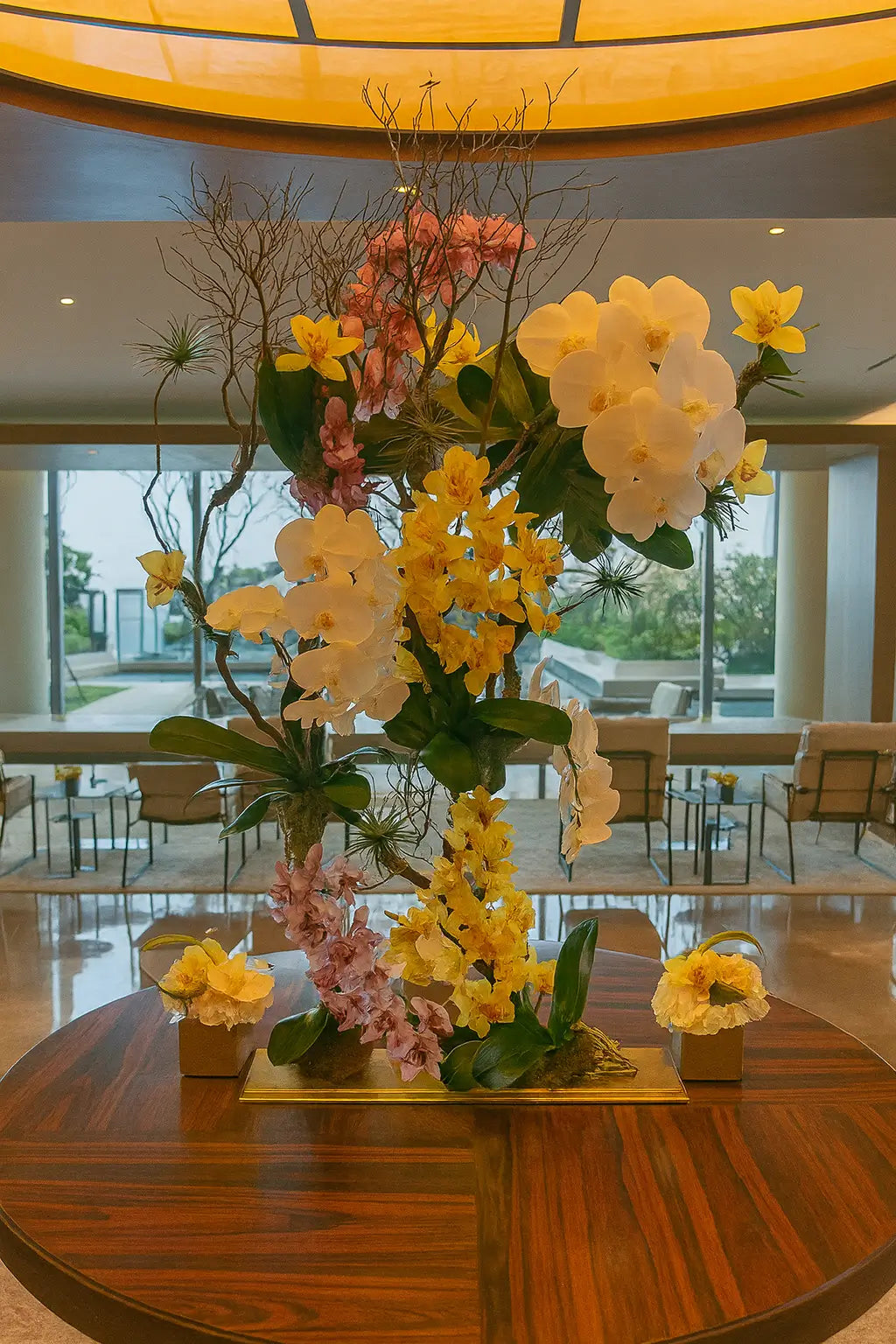A large luxurious floral arrangement of white, yellow and pink orchids on an elegant metal stand, placed in the three-story glass lobby of Turnberry Ocean Club Residences overlooking the sea horizon and a contemporary interior.