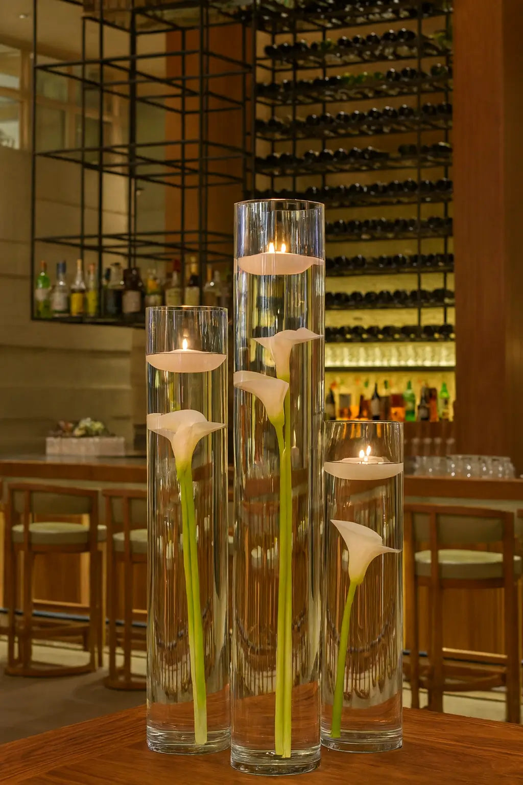 A bright daytime view of Zuma Miami’s bar area with high ceilings, a wine wall in the background, and tall glass vases filled with floating candles and white calla lilies, creating a luxurious lounge atmosphere.