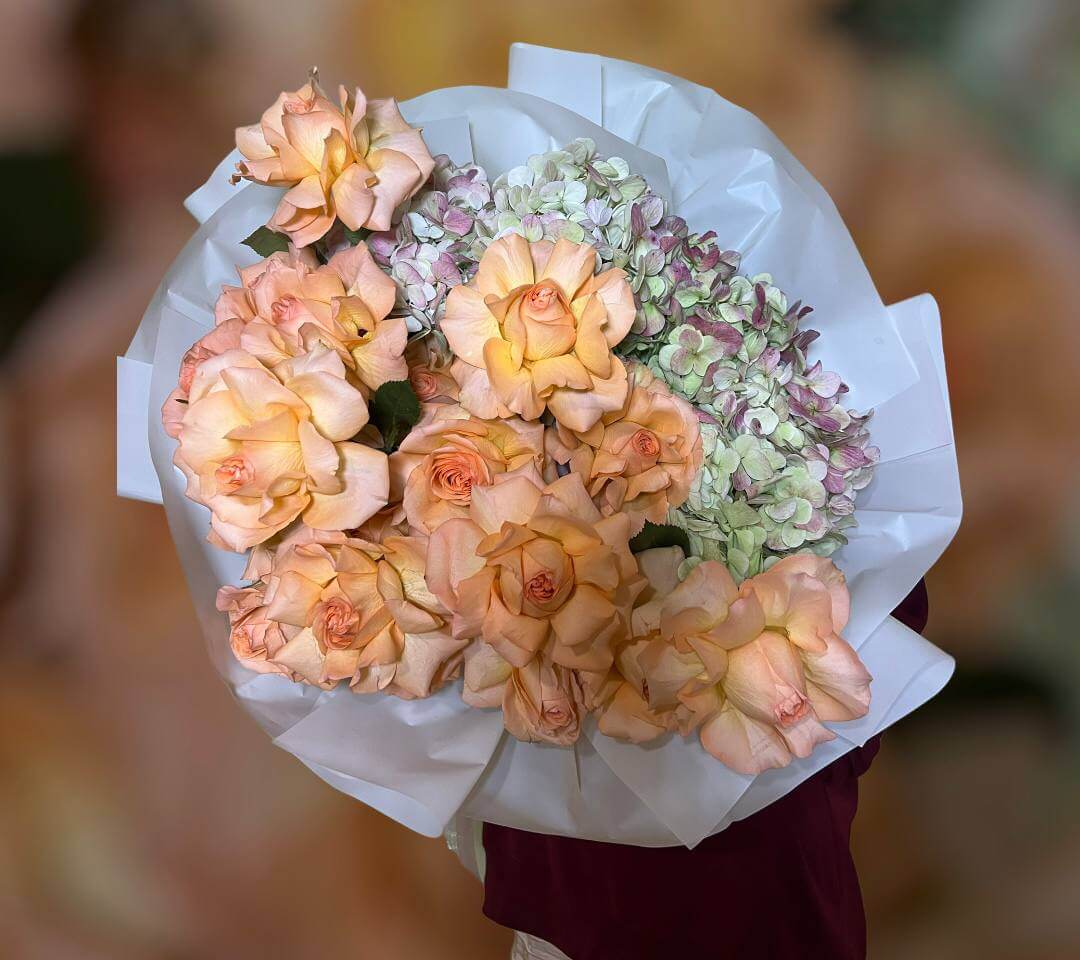 Orange Mood bouquet with peach roses and pastel hydrangeas in white wrap held by elegant woman — luxury floral Miami