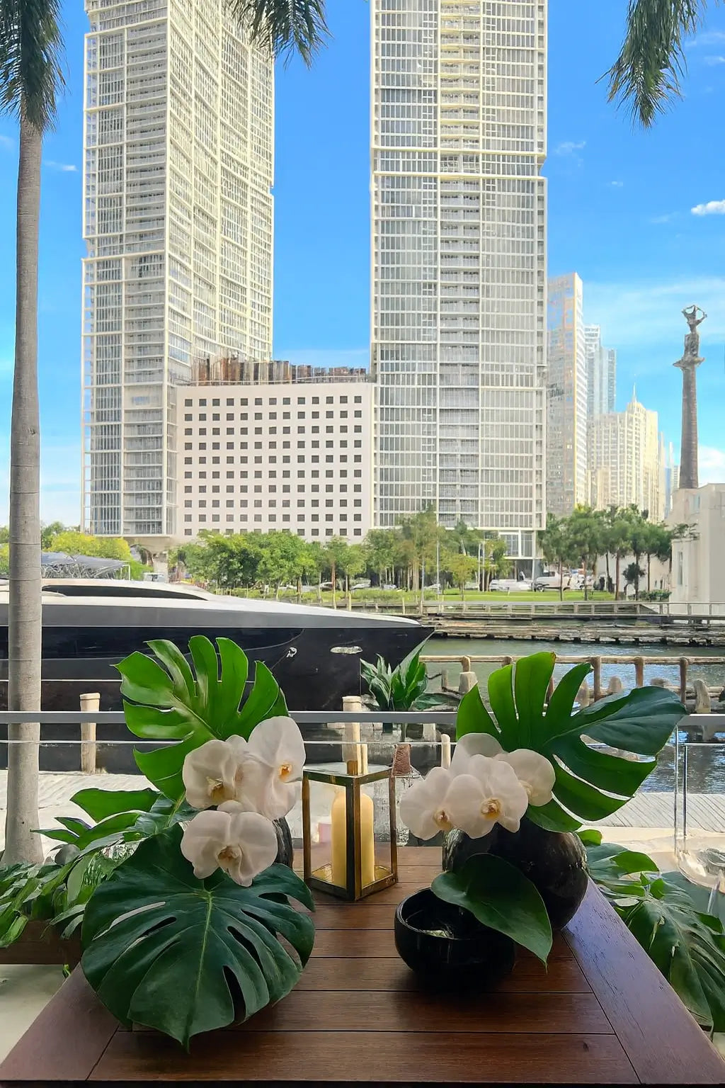 A terrace at Zuma Miami overlooking the skyscrapers and water, with low Japanese lacquer vases holding white orchids and monstera greenery, softly lit by lanterns. The scene conveys a relaxed dining atmosphere with an exclusive panoramic view.