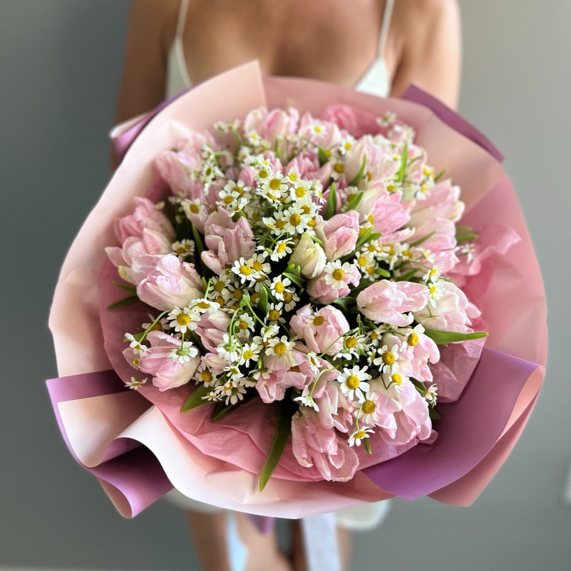 Hand-tied bouquet with pink tulips and chamomile wrapped in pink and violet paper held by woman in garden — unforgettable flower gift