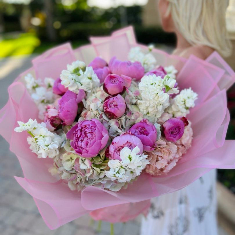 A young blonde woman in a white luxury dress gently smiles while holding a lush bouquet of pink peonies and white stock flowers