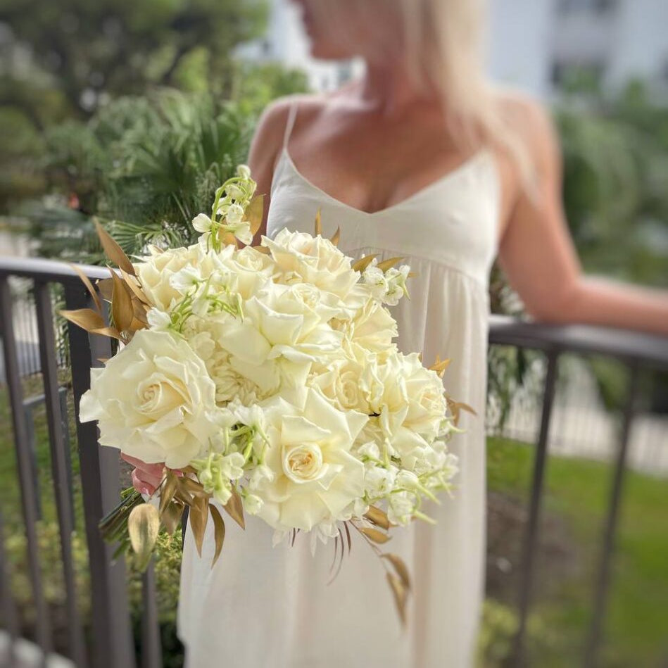 Elegant bouquet with white roses and golden leaves held by woman on balcony — luxury hand-tied flower delivery in Miami